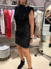 Load image into Gallery viewer, A woman modeling a sleeveless black sequined mini dress in a boutique setting, standing in front of a clothing rack and mirror.
